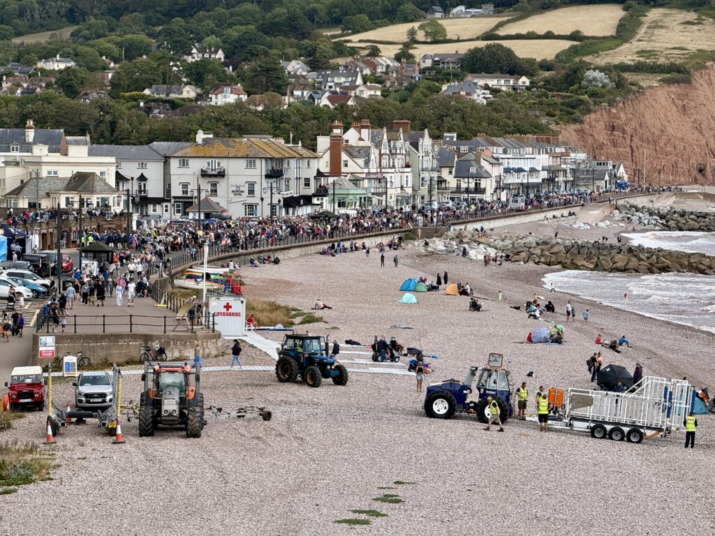 Crowds along Sidmouth seafront and pebble beach in Devon during the Regatta weekend, with event vehicles on the beach and red cliffs beyond.