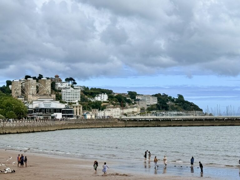 Visitors walking and paddling on Torre Abbey Sands, with Torquay seafront, Princess Theatre, marina and hillside buildings beyond.