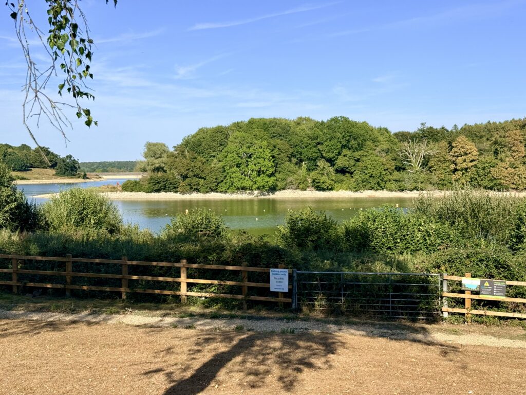  View across Sutton Bingham Reservoir from a gated area marked for fishing permit holders only, with low water exposing the shoreline beneath wooded banks.