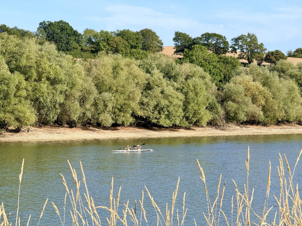 Two people paddling a tandem kayak across Sutton Bingham Reservoir, with wooded countryside beyond.