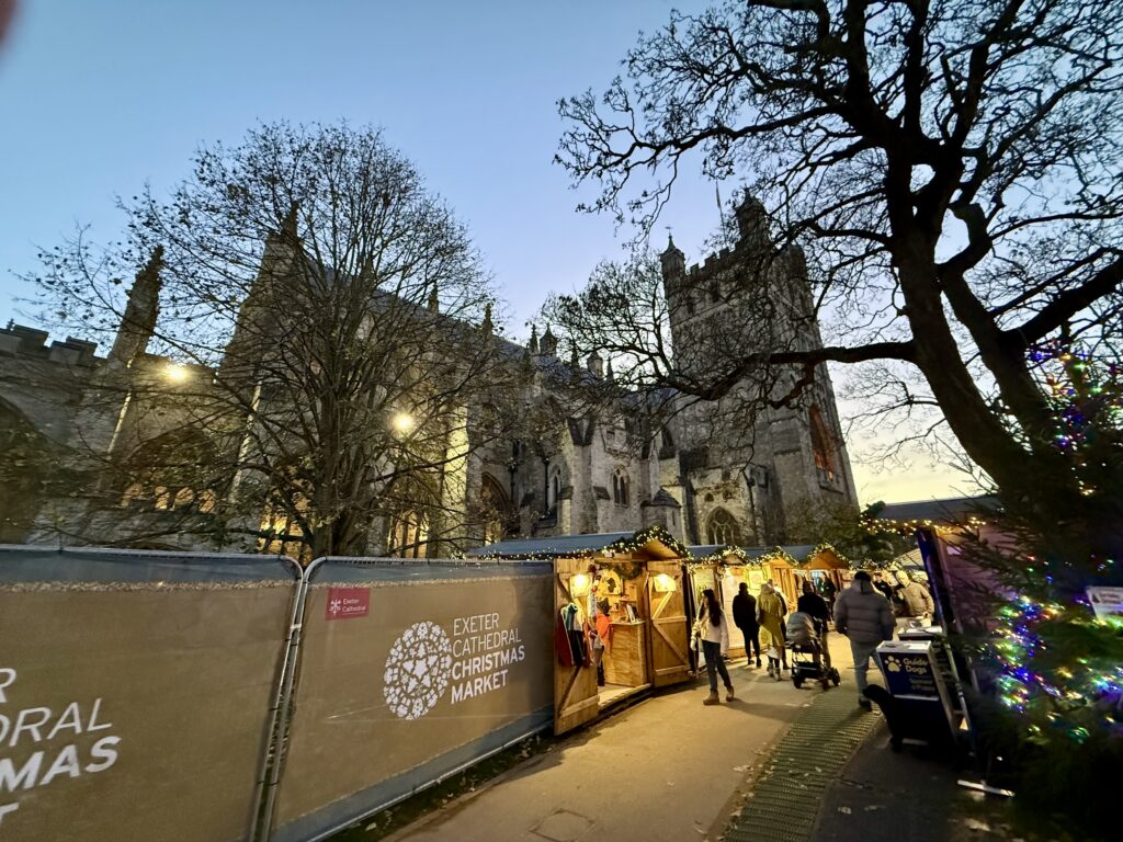 Visitors browsing illuminated Christmas Market chalets beside Exeter Cathedral at dusk