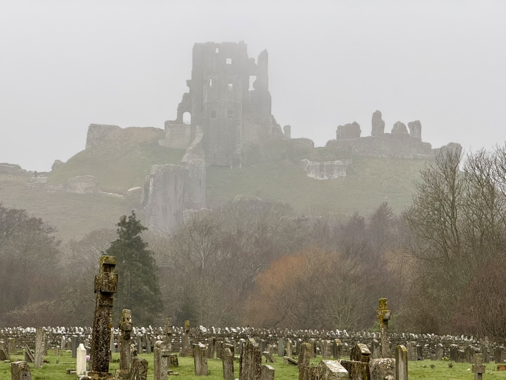 Corfe Castle ruins rising through mist above gravestones in the village churchyard.
