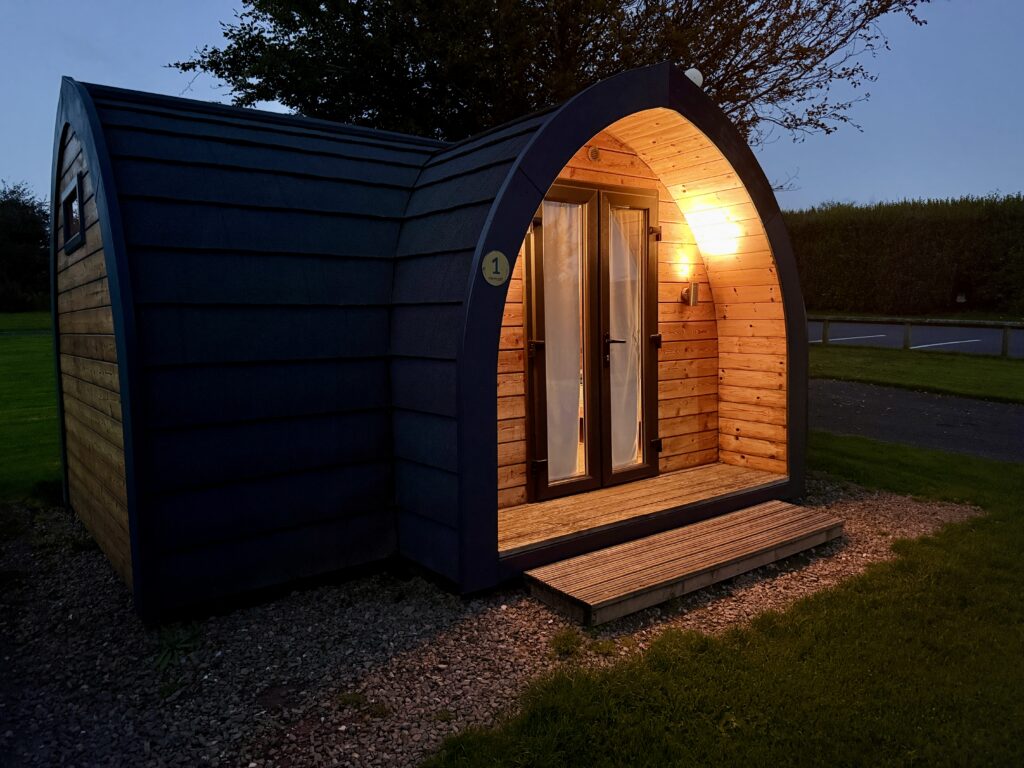 Camping pod at Stowford Farm Meadows illuminated at night, with wooden entrance, double doors and small deck.