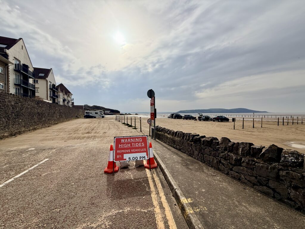 Royal Sands at Weston-super-Mare with cars parked on the sand, a high-tide warning sign and Brean Down visible across the bay.