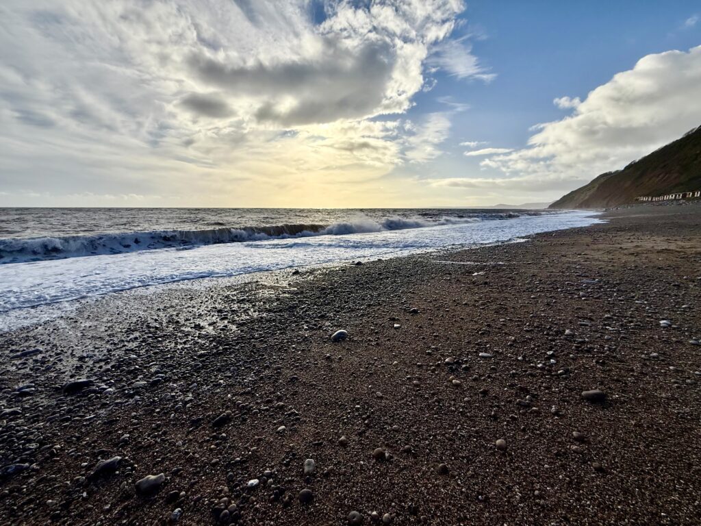 Waves breaking across the broad pebble beach at Branscombe beneath dramatic clouds, with steep cliffs stretching along the East Devon coast.