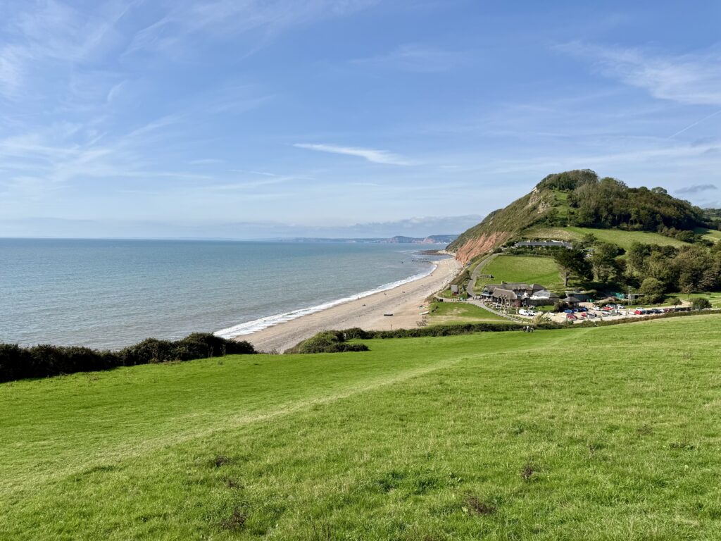View across green fields to Branscombe Beach, the Sea Shanty café and wooded cliffs on the Jurassic Coast in Devon.