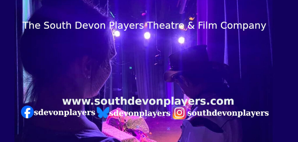 South Devon Players Theatre & Film Company promotional banner showing a theatre scene under purple stage lighting, with website and social media details.