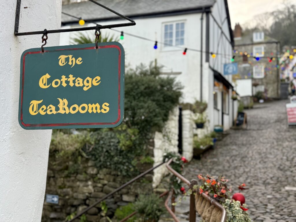 Cottage Tea Rooms sign beside Clovelly's narrow cobbled street, with whitewashed cottages beyond.