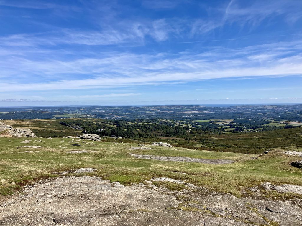 Panoramic view from Haytor across Dartmoor towards the rolling Devon countryside and distant coast beneath a blue summer sky.