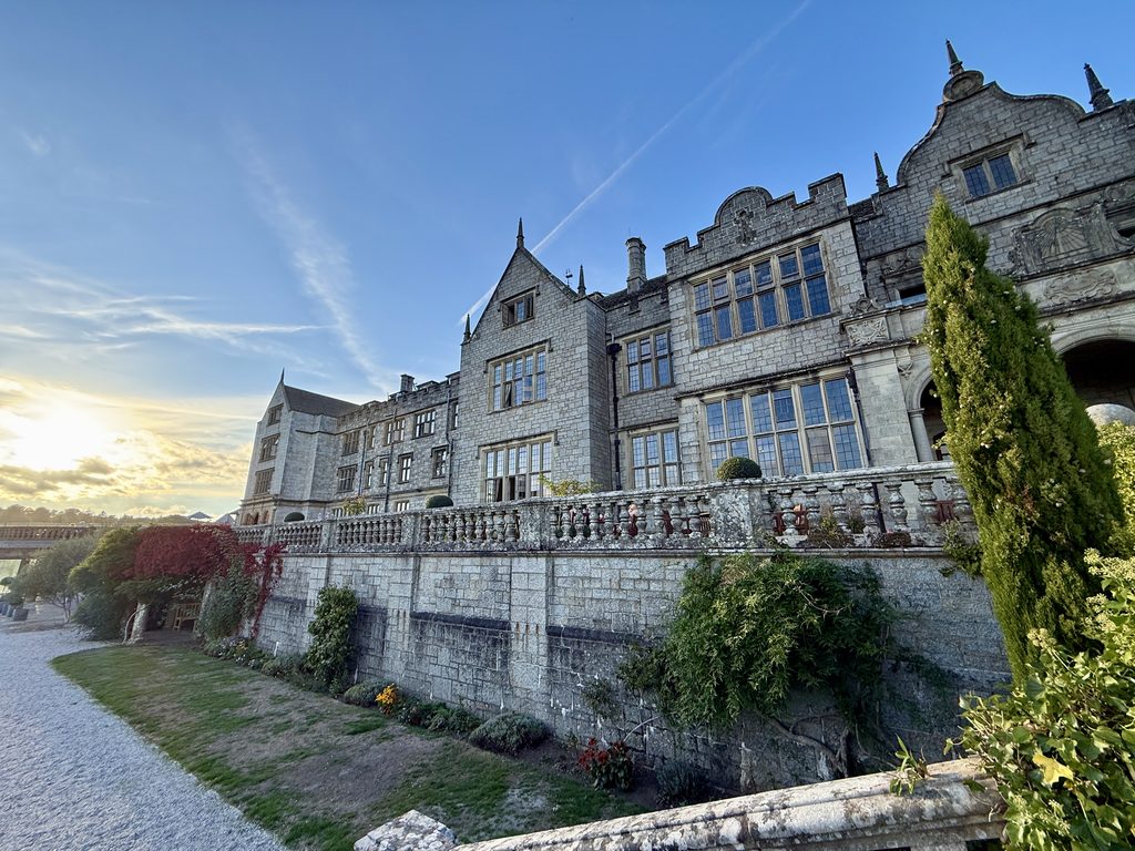 Wide view of Bovey Castle’s grand stone façade and terrace, with formal gardens beneath and evening sunlight over the Dartmoor estate in Devon.