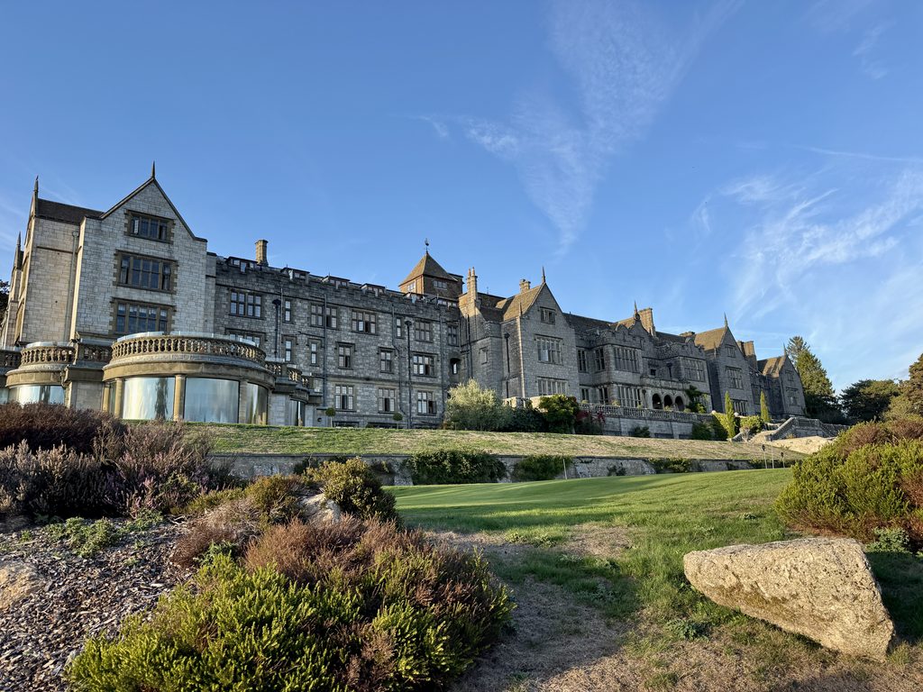 Wide view of Bovey Castle from its landscaped grounds, showing the historic stone hotel, terraces, lawns and gardens beneath a blue Dartmoor sky.