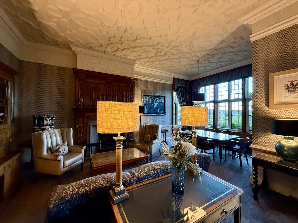 Elegant lounge at Bovey Castle with an ornate plaster ceiling, carved wooden fireplace, armchairs, table lamps and tall windows overlooking the Devon countryside.