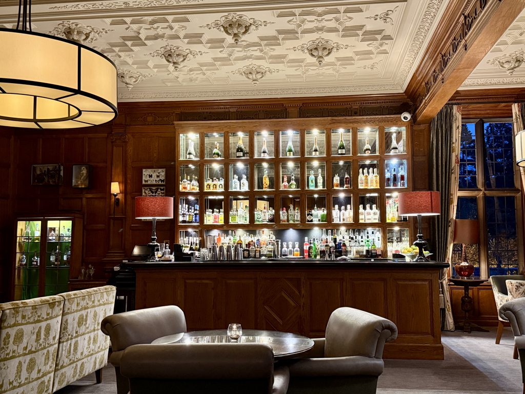 The Oak Bar at Bovey Castle, with an illuminated drinks display, carved wood panelling, ornate plaster ceiling and comfortable lounge seating.