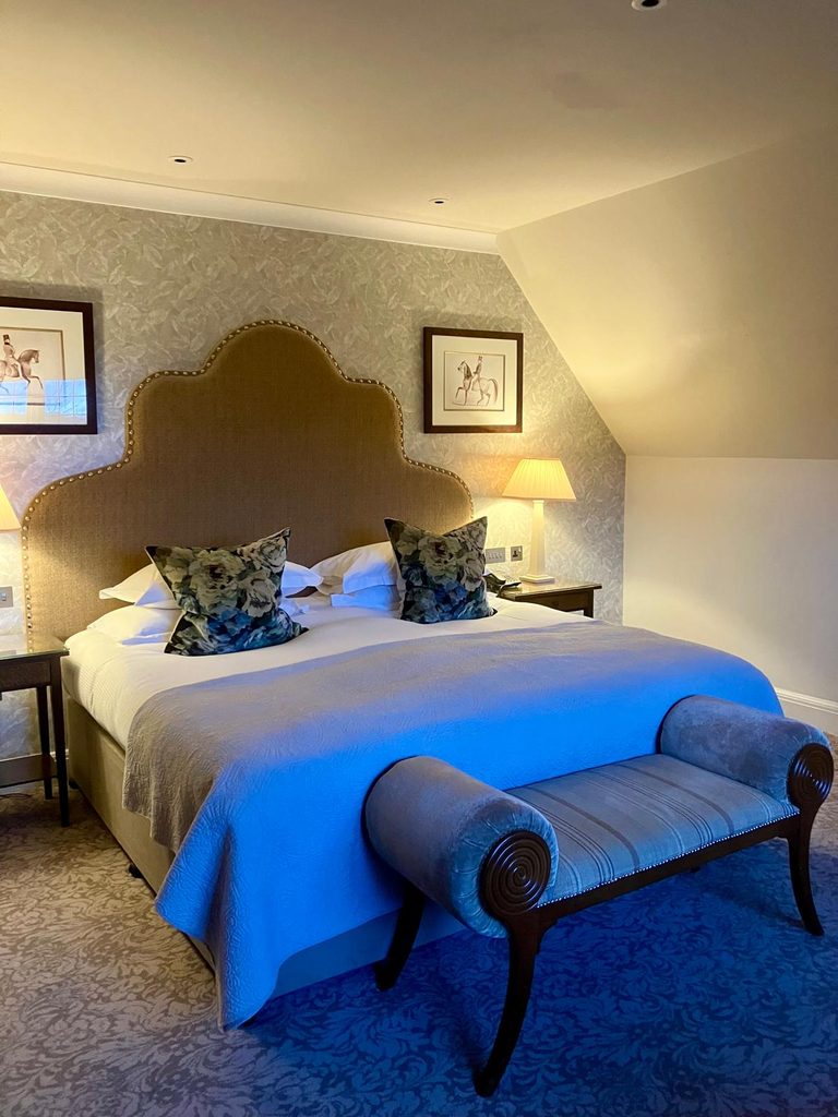 Bedroom at Bovey Castle with a large double bed, upholstered headboard, floral cushions, bedside lamps and a blue bench at the foot of the bed.