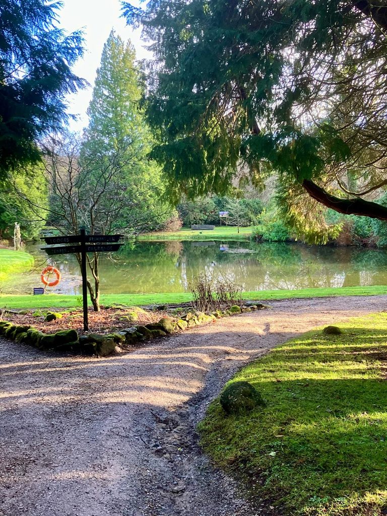Woodland path beside a lake in the grounds of Bovey Castle, with a signpost pointing towards the lodges, fishing lake, tennis courts and driving range.