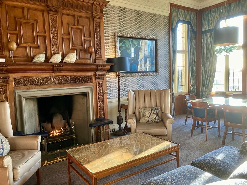 Elegant lounge at Bovey Castle with a lit fireplace, ornate carved woodwork, armchairs and tall windows overlooking the Dartmoor grounds.